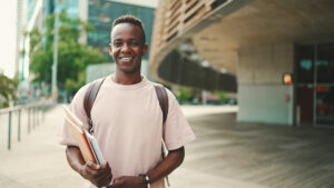 Young african student stands outside of university, looks at the camera, smiles